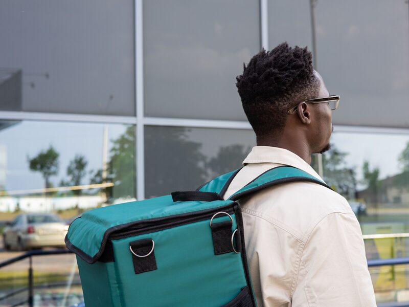 A delivery rider in glasses and a cream shirt carrying a green insulated food delivery bag on his back