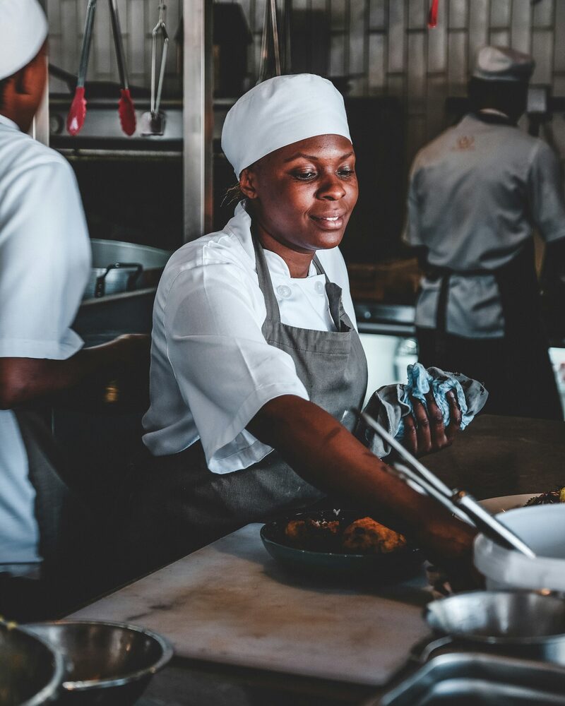A smiling cook in a white cap and grey apron plating food in a busy kitchen